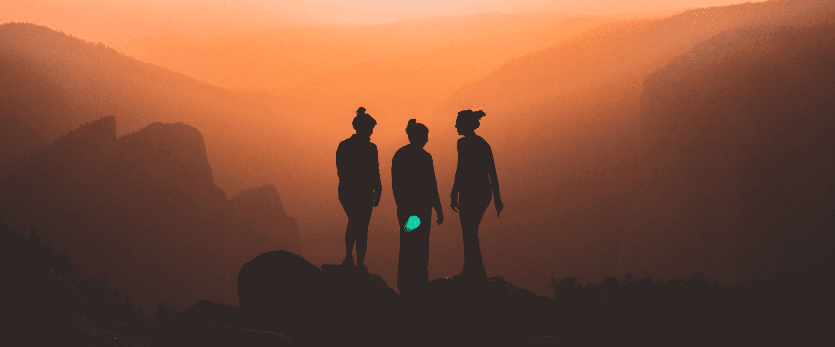An image of three women standing on a high point and looking out at mountains and formations with a hazy and burnt orange sky.